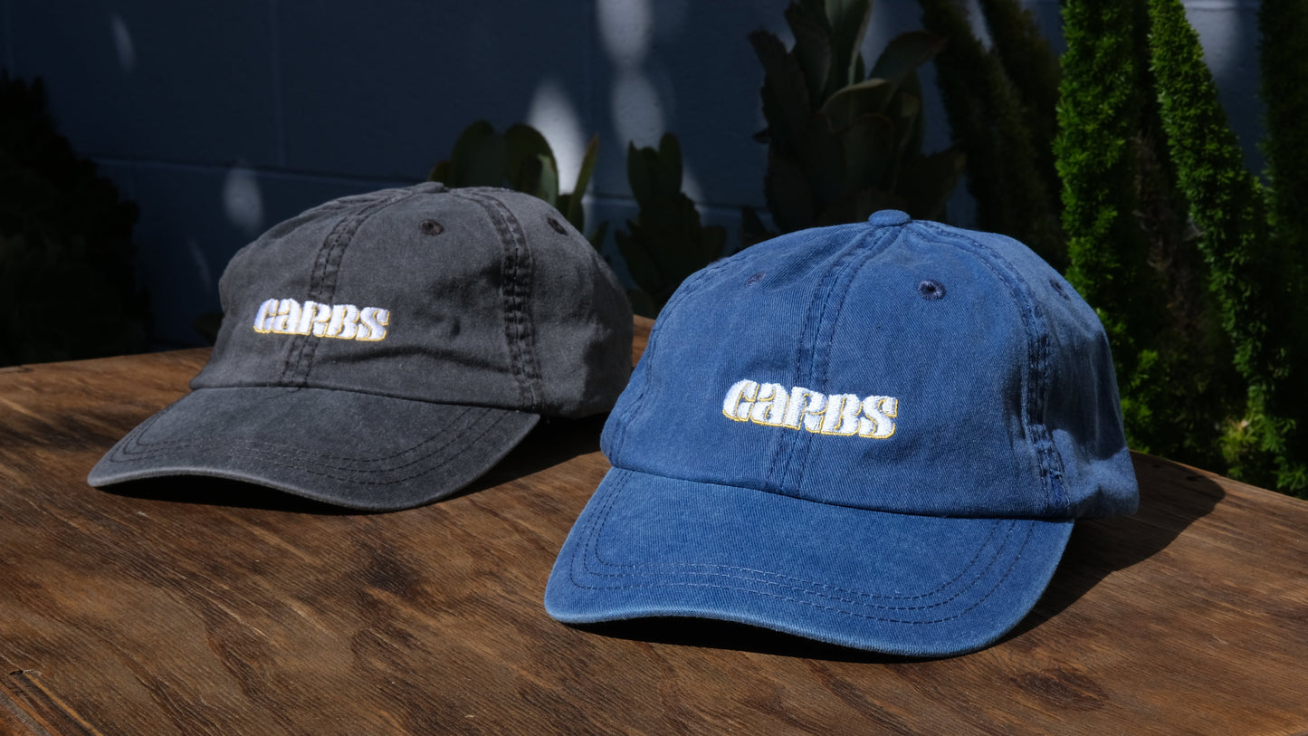 Two baseball caps, one gray and one blue, on a wooden surface with a blurred outdoor background.